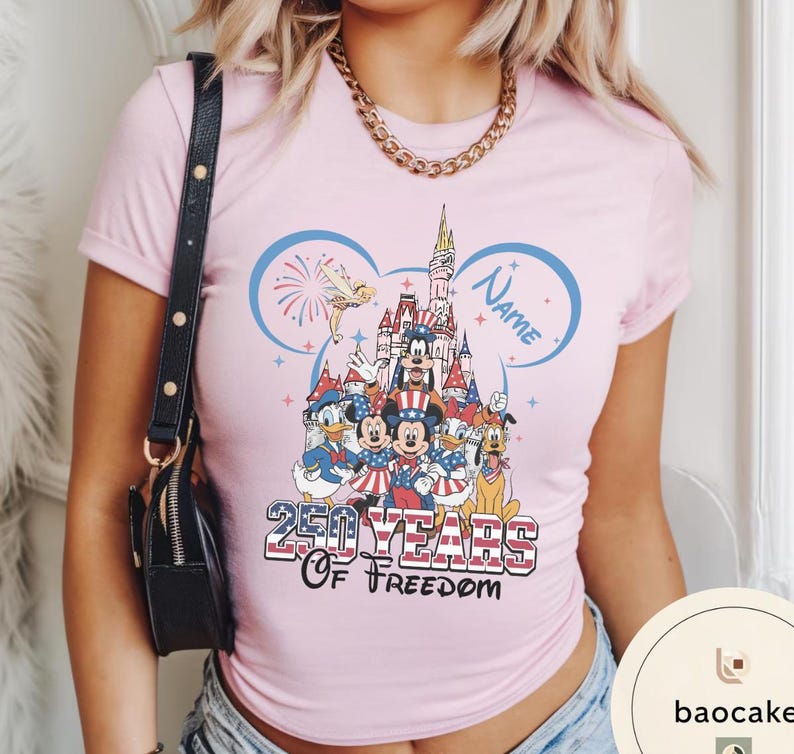 Peut inclure: T-shirt rose clair avec un motif de personnages Disney en tenues patriotiques devant un ch&acirc;teau. Le t-shirt porte l'inscription "250 Years of Freedom" et un espace pour un nom dans un contour d'oreille de Mickey.