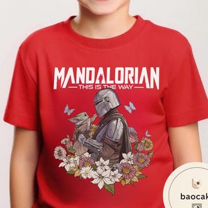 Puede incluir: Camiseta roja con un dise&ntilde;o de The Mandalorian y Grogu rodeados de flores y mariposas. El texto "MANDALORIAN - THIS IS THE WAY" est&aacute; impreso en blanco en la parte superior.