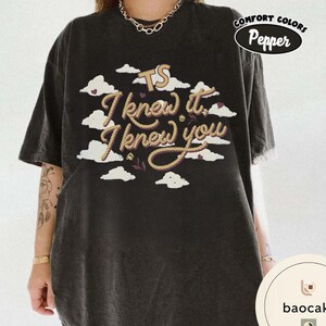 K&ouml;nnte beinhalten: Ein anthrazitfarbenes Kurzarm-T-Shirt mit einem Grafikdruck aus wei&szlig;en Wolken, kleinen Herzen und dem Text TS I knew it, I knew you in einer seilartigen Schriftart. Das T-Shirt hat ein Comfort Colors Pepper-Etikett in der oberen rechten Ecke.