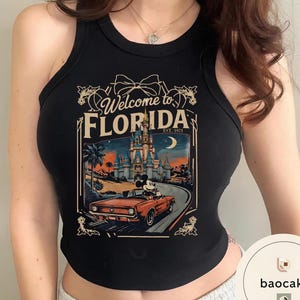 May include: Black tank top with a graphic featuring a vintage Florida travel design. The design includes a castle, Mickey Mouse in a red convertible, and the text "Welcome to Florida, Est. 1971."