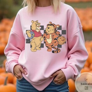 May include: Pink sweatshirt featuring a cartoon illustration of Winnie the Pooh and Tigger holding Halloween pumpkins. The characters stand in front of a black and white checkered background. The sweatshirt is a solid pink color.