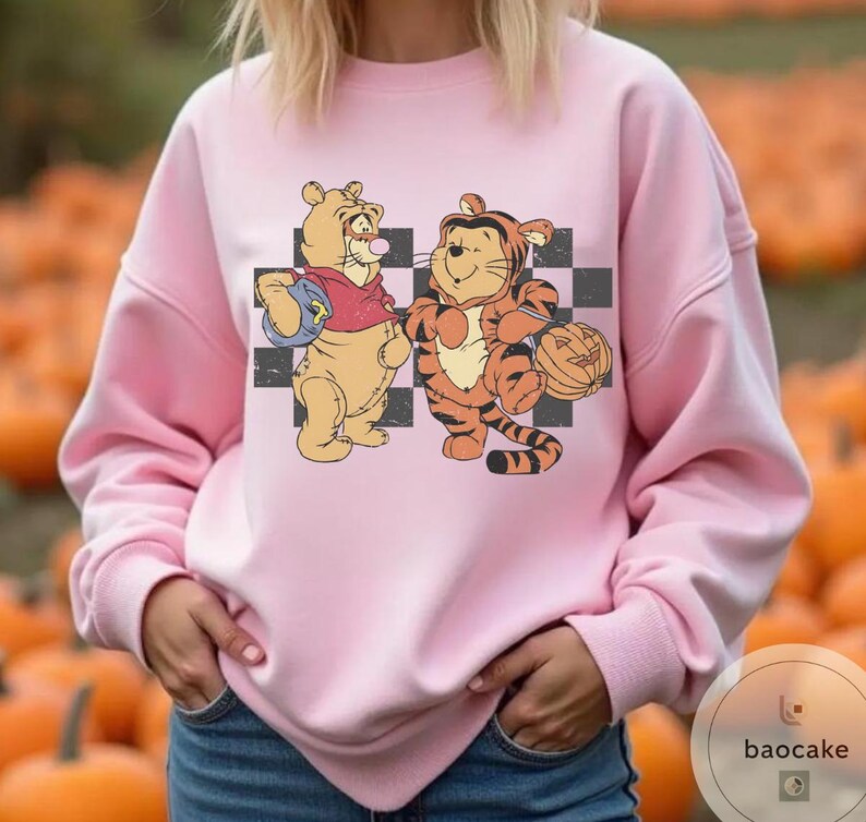 May include: Pink sweatshirt featuring a cartoon illustration of Winnie the Pooh and Tigger holding Halloween pumpkins. The characters stand in front of a black and white checkered background. The sweatshirt is a solid pink color.