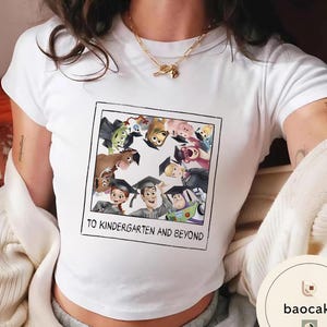 May include: White t-shirt with a colourful graphic of Toy Story characters wearing graduation caps. The image is framed with a black border and includes the text "TO KINDERGARTEN AND BEYOND".