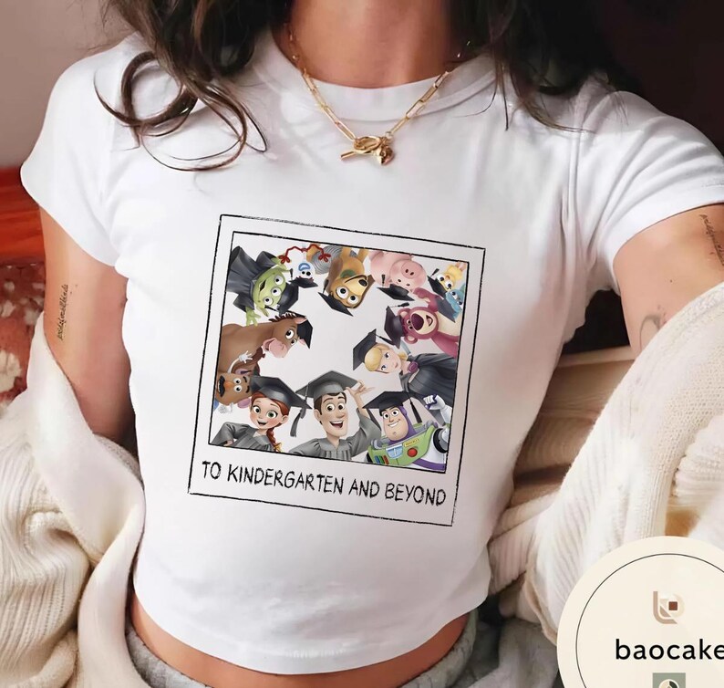 May include: White t-shirt with a colourful graphic of Toy Story characters wearing graduation caps. The image is framed with a black border and includes the text "TO KINDERGARTEN AND BEYOND".
