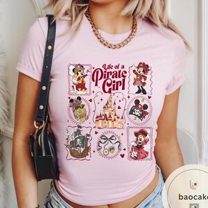 May include: Light pink t-shirt featuring a graphic design with the text "Life of a Pirate Girl." The design includes various illustrations of a mouse character in pirate attire, a pirate ship, and a castle.