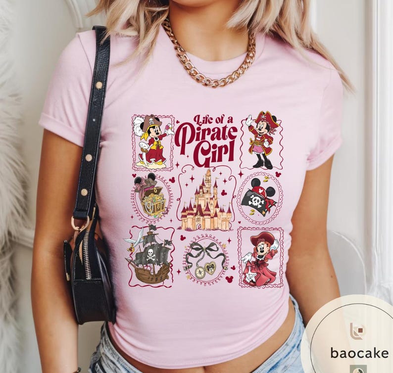 May include: Light pink t-shirt featuring a graphic design with the text "Life of a Pirate Girl." The design includes various illustrations of a mouse character in pirate attire, a pirate ship, and a castle.