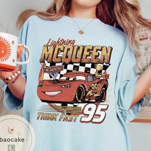 Puede incluir: Camiseta azul claro con un dise&ntilde;o de Rayo McQueen de la pel&iacute;cula Cars. El dise&ntilde;o incluye el texto "Lightning McQueen", "Cars", "Think Fast" y el n&uacute;mero "95". La camiseta tiene un aspecto vintage y desgastado.
