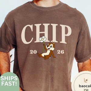 May include: Brown t-shirt featuring the word "CHIP" in cream-colored block letters, with the numbers "20" and "26" on either side of a cartoon chipmunk holding a flower. The shirt has a crew neck and short sleeves.