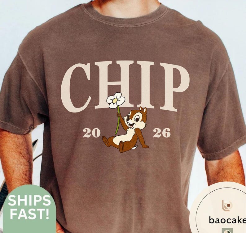 May include: Brown t-shirt featuring the word "CHIP" in cream-colored block letters, with the numbers "20" and "26" on either side of a cartoon chipmunk holding a flower. The shirt has a crew neck and short sleeves.
