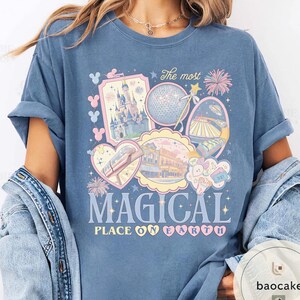 May include: A blue t-shirt with a colourful graphic featuring theme park imagery, including a castle, a geodesic sphere, and a monorail. The text "The most MAGICAL PLACE ON EARTH" is displayed in a whimsical font. The shirt has rolled-up sleeves.