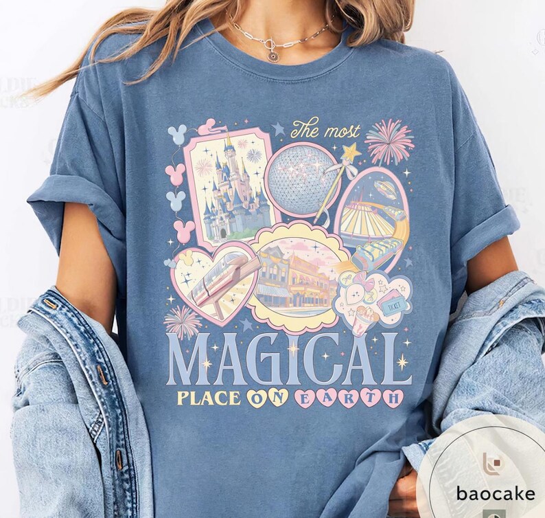 May include: A blue t-shirt with a colourful graphic featuring theme park imagery, including a castle, a geodesic sphere, and a monorail. The text "The most MAGICAL PLACE ON EARTH" is displayed in a whimsical font. The shirt has rolled-up sleeves.