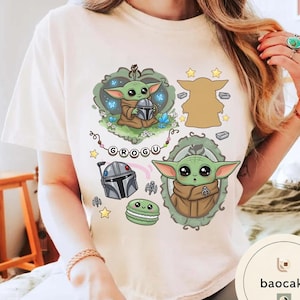 May include: A cream-colored t-shirt featuring a graphic print of the character Grogu. The design includes Grogu holding a helmet, a silhouette, a framed portrait, a Mandalorian helmet, a green macaron, stars, and the text GROGU on a banner.