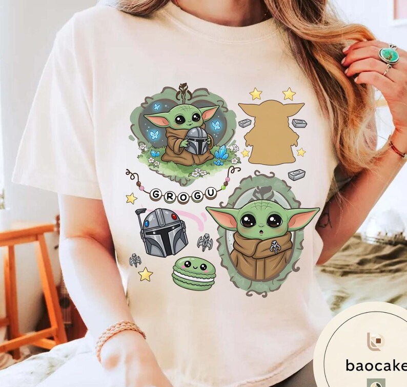 May include: A cream-colored t-shirt featuring a graphic print of the character Grogu. The design includes Grogu holding a helmet, a silhouette, a framed portrait, a Mandalorian helmet, a green macaron, stars, and the text GROGU on a banner.
