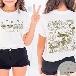 May include: White t-shirts with green and brown illustrations. One shirt features "Animal Kingdom" text and cartoon images. The other shirt has "Kilimanjaro" text and a detailed adventure scene with a hot air balloon and characters.