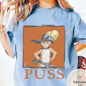 May include: Light blue t-shirt featuring a cartoon illustration of a cat in a hat and cape, with the word "PUSS" in large letters below the image. The cat is orange and white, with a black hat and cape.