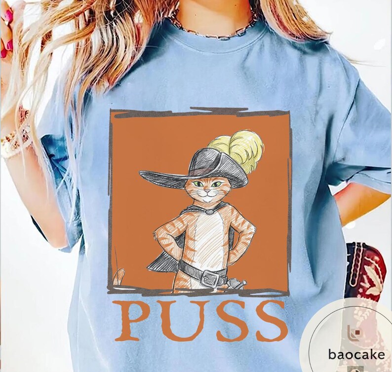 May include: Light blue t-shirt featuring a cartoon illustration of a cat in a hat and cape, with the word "PUSS" in large letters below the image. The cat is orange and white, with a black hat and cape.