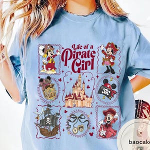 May include: Light blue t-shirt featuring a design with the text "Life of a Pirate Girl." The design includes illustrations of Minnie Mouse in pirate attire, a treasure chest, a pirate ship, and a castle.