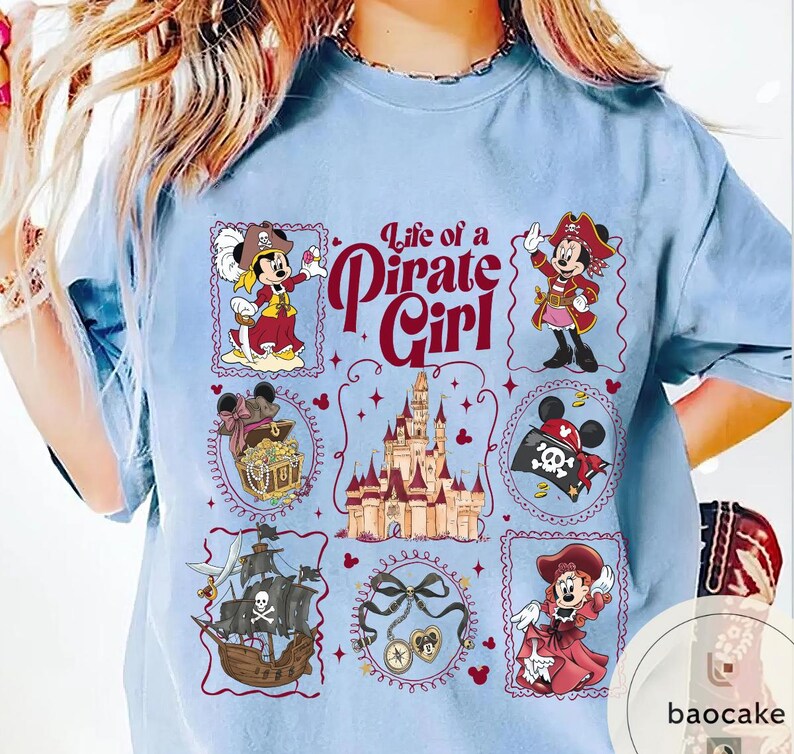 May include: Light blue t-shirt featuring a design with the text "Life of a Pirate Girl." The design includes illustrations of Minnie Mouse in pirate attire, a treasure chest, a pirate ship, and a castle.