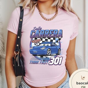 Puede incluir: Camiseta rosa claro con un dise&ntilde;o de Sally Carrera de la pel&iacute;cula Cars. El dise&ntilde;o incluye el texto "Sally Carrera", "Cars" y "Think Fast 301" con una ilustraci&oacute;n de un coche azul.