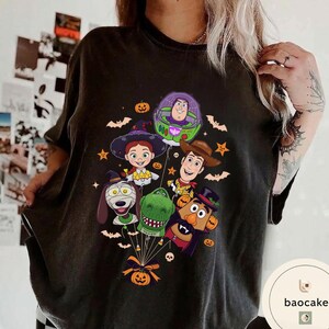 Pode incluir: Uma t-shirt preta de manga curta com uma impress&atilde;o gr&aacute;fica de personagens de Toy Story vestidos com fantasias de Halloween, incluindo Buzz Lightyear, Woody, Jessie, Rex, Slinky Dog e Mr. Potato Head, todos ligados por fios a uma ab&oacute;bora.