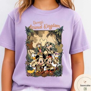 May include: A light purple t-shirt featuring a graphic of Disney's Animal Kingdom characters, including Mickey and Minnie Mouse, Donald Duck, and Goofy. The design is set within a jungle-themed frame with the text "Disney's Animal Kingdom".