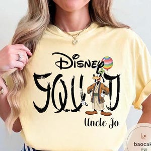 Puede incluir: Camiseta amarilla clara con la inscripci&oacute;n "Disney Goofy Uncle Jo" en escritura negra. El dise&ntilde;o incluye una imagen de dibujos animados de Goofy sosteniendo un globo de arco&iacute;ris. La camiseta tiene mangas cortas.