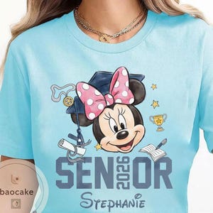 May include: Light blue t-shirt featuring Minnie Mouse wearing a graduation cap and a pink polka dot bow. The shirt has the text "Senior 2026" and the name "Stephanie" in a script font. Graduation-themed design.