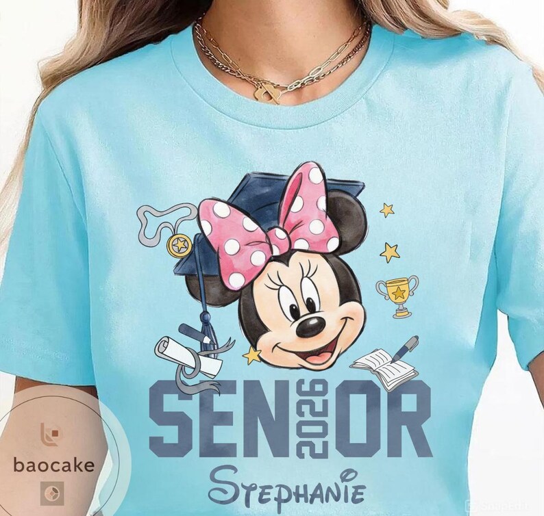 May include: Light blue t-shirt featuring Minnie Mouse wearing a graduation cap and a pink polka dot bow. The shirt has the text "Senior 2026" and the name "Stephanie" in a script font. Graduation-themed design.