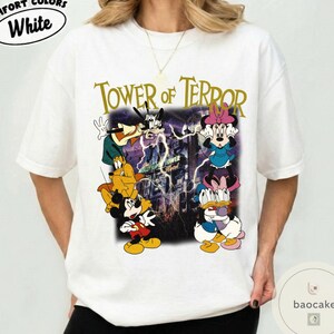 May include: White t-shirt featuring a graphic of Disney characters, including Mickey Mouse, Goofy, and Minnie Mouse, with the text "Tower of Terror." The design has a vintage, distressed look with a purple and yellow color scheme.