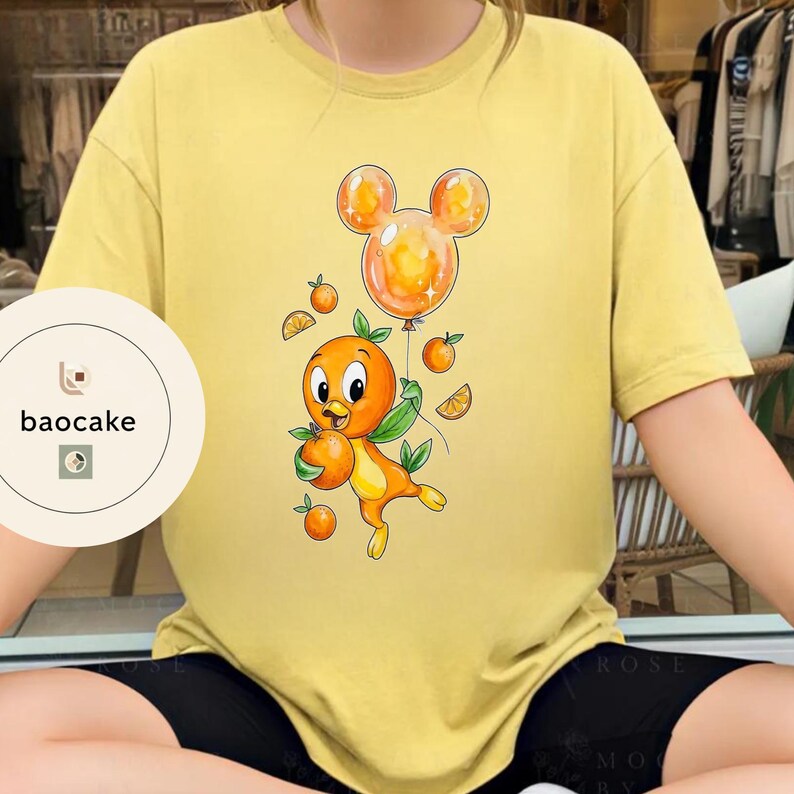 Op de afbeelding: Geel T-shirt met een cartoon oranje vogel die sinaasappels en een Mickey Mouse-vormige ballon vasthoudt. De vogel is oranje en geel met groene bladeren. De ballon is oranje. Het shirt heeft het logo "baocake" links.