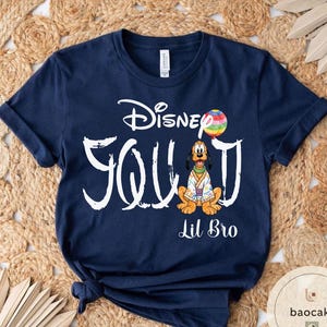 Puede incluir: Camiseta azul marino con la inscripci&oacute;n blanca "Disney Squad" y "Lil Bro". El dise&ntilde;o incluye un personaje de dibujos animados de Pluto con un atuendo colorido y un globo arco&iacute;ris.
