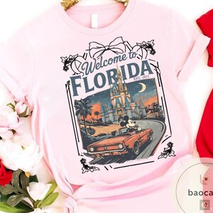 May include: Light pink t-shirt featuring a graphic of Mickey Mouse driving a red convertible in front of a castle, with the text "Welcome to Florida" and "Est. 1971". The design includes palm trees and a crescent moon.