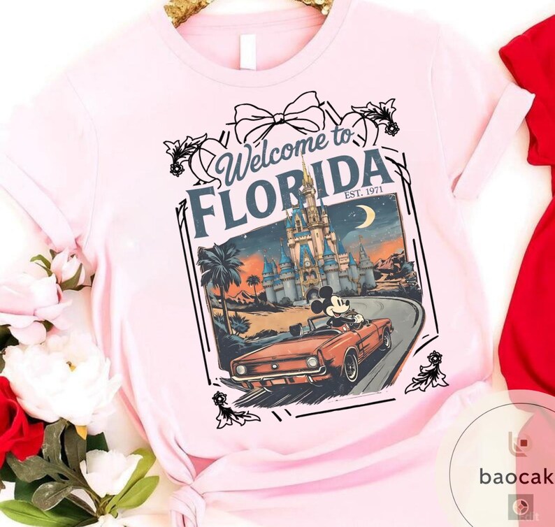 May include: Light pink t-shirt featuring a graphic of Mickey Mouse driving a red convertible in front of a castle, with the text "Welcome to Florida" and "Est. 1971". The design includes palm trees and a crescent moon.