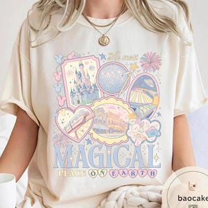 May include: Cream-coloured t-shirt with a pastel graphic featuring a castle, Spaceship Earth, and other theme park imagery. The text "MAGICAL PLACE ON EARTH" is displayed in blue and pink lettering, creating a whimsical aesthetic.
