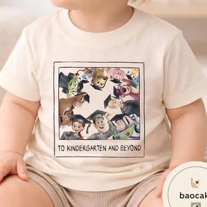 May include: Cream-coloured t-shirt featuring a framed graphic of animated characters wearing graduation caps. The graphic includes the text "TO KINDERGARTEN AND BEYOND". The shirt is made of a soft material.