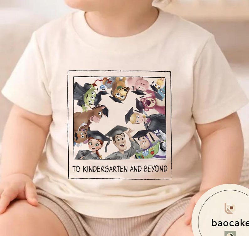 May include: Cream-coloured t-shirt featuring a framed graphic of animated characters wearing graduation caps. The graphic includes the text "TO KINDERGARTEN AND BEYOND". The shirt is made of a soft material.