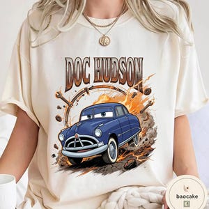 May include: Cream-coloured t-shirt with a cartoon illustration of Doc Hudson, a blue car with white-walled tyres, from the film Cars. The text "DOC HUDSON" is above the car in brown lettering. The design includes a clock, spanners, and flames.