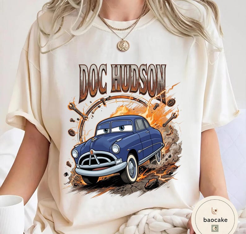May include: Cream-coloured t-shirt with a cartoon illustration of Doc Hudson, a blue car with white-walled tyres, from the film Cars. The text "DOC HUDSON" is above the car in brown lettering. The design includes a clock, spanners, and flames.