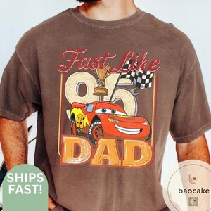 May include: Brown t-shirt featuring a graphic of the character Lightning McQueen from the movie Cars, with the text "Fast Like Dad." The design includes a trophy, checkered flag, and the number 95. The word "DAD" is in large orange letters.