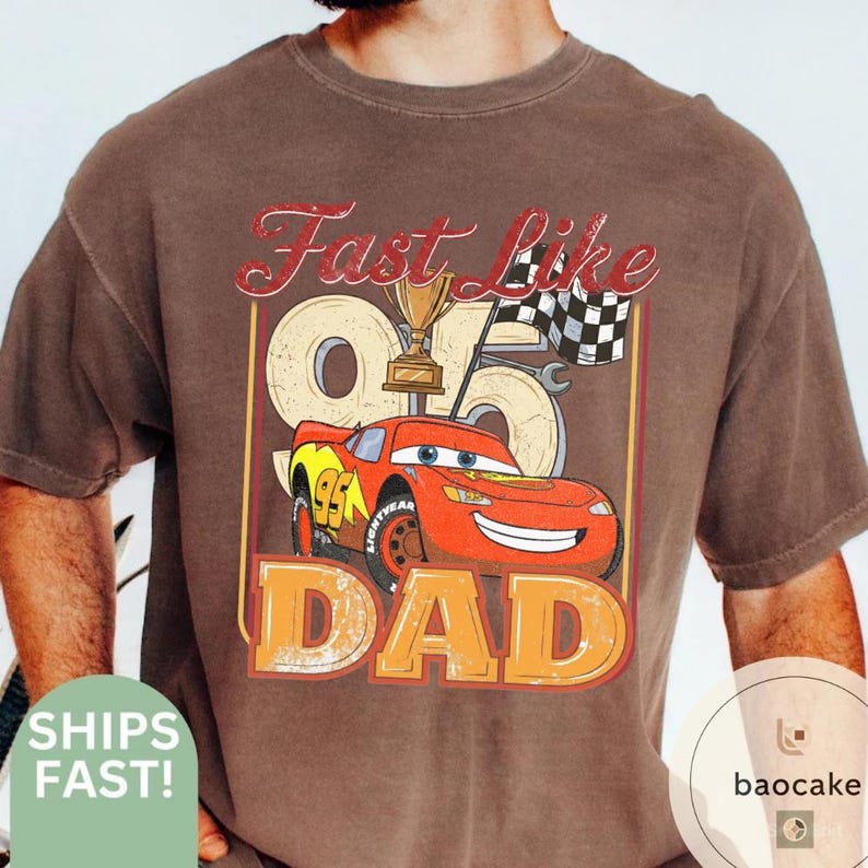 May include: Brown t-shirt featuring a graphic of the character Lightning McQueen from the movie Cars, with the text "Fast Like Dad." The design includes a trophy, checkered flag, and the number 95. The word "DAD" is in large orange letters.