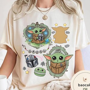 May include: A cream-colored t-shirt featuring a graphic print of the character Grogu in various poses, a silver helmet, a green macaron, and the text "GROGU" on a string of beads, surrounded by small stars, blue butterflies, and crystals.