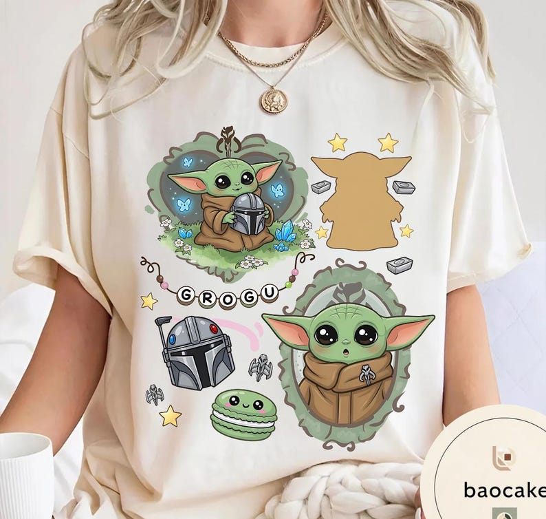 May include: A cream-colored t-shirt featuring a graphic print of the character Grogu in various poses, a silver helmet, a green macaron, and the text "GROGU" on a string of beads, surrounded by small stars, blue butterflies, and crystals.