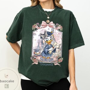 May include: Dark green t-shirt featuring a framed graphic of Daisy Duck in a graduation gown, holding a diploma. The graphic includes a bow, flowers, fireworks, and the text "SENIOR Stephanie".