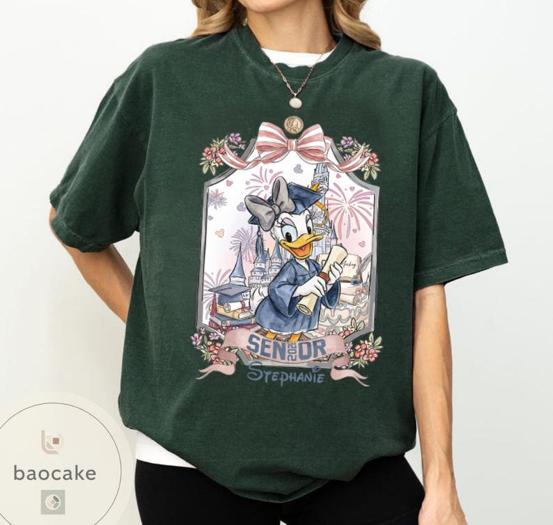 May include: Dark green t-shirt featuring a framed graphic of Daisy Duck in a graduation gown, holding a diploma. The graphic includes a bow, flowers, fireworks, and the text "SENIOR Stephanie".