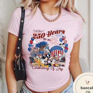 Puede incluir: Camiseta rosa claro con un dise&ntilde;o de Mickey, Minnie, Donald y Goofy celebrando. El dise&ntilde;o incluye el texto "Oh Boy! 250 Years Celebrating of Joy" con fuegos artificiales, globos y banderas americanas.