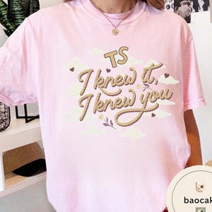 K&ouml;nnte beinhalten: Ein hellrosa Kurzarm-T-Shirt mit einem Grafikdruck, der den Text TS I knew it, I knew you in einer seilartigen Schrift zeigt. Das Design enth&auml;lt cremefarbene Wolken, kleine dunkelbraune Herzen und florale Akzente um den Schriftzug.