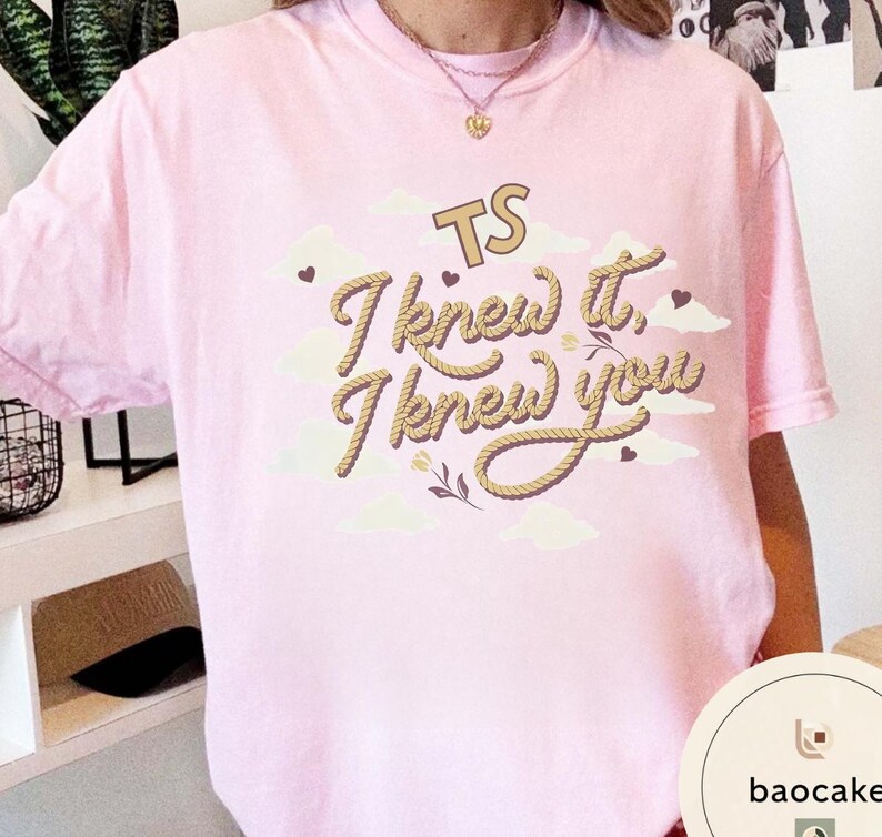 K&ouml;nnte beinhalten: Ein hellrosa Kurzarm-T-Shirt mit einem Grafikdruck, der den Text TS I knew it, I knew you in einer seilartigen Schrift zeigt. Das Design enth&auml;lt cremefarbene Wolken, kleine dunkelbraune Herzen und florale Akzente um den Schriftzug.