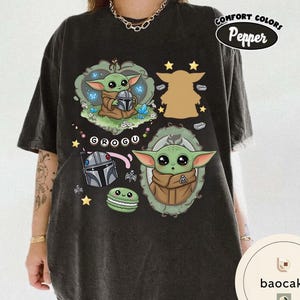 May include: A charcoal gray t-shirt featuring a graphic print of Grogu in two ornate frames, a Mandalorian helmet, a green macaron, and the text "GROGU". The design includes small stars and blue butterflies, with a "Comfort Colors Pepper" label.