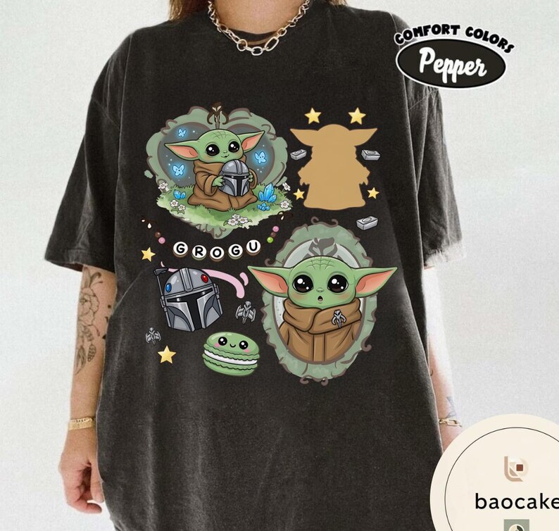 May include: A charcoal gray t-shirt featuring a graphic print of Grogu in two ornate frames, a Mandalorian helmet, a green macaron, and the text "GROGU". The design includes small stars and blue butterflies, with a "Comfort Colors Pepper" label.