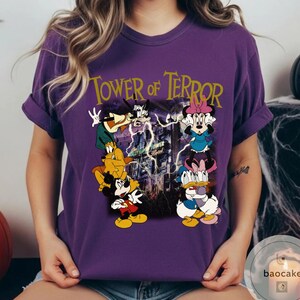 May include: Purple t-shirt featuring a graphic of Disney characters, including Mickey Mouse, Goofy, Minnie Mouse, and Donald Duck, with the text "Tower of Terror" in gold lettering. The design has a spooky, Halloween theme.